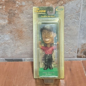 Puede incluir: Una figura de Tiger Woods con cabeza oscilante en un embalaje de plástico transparente. La figura lleva una camisa roja y pantalones negros. El embalaje dice "THE MAJORS PlayMakers" y "Collect All Four!"