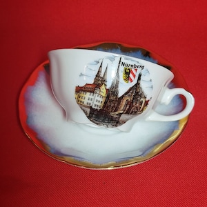 May include: White porcelain teacup and saucer with a gold rim. The teacup features a colorful illustration of the Frauenkirche church in Nuremberg, Germany. The saucer has a blue and white swirl design.