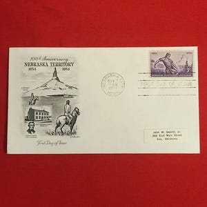 May include: A white envelope with a Nebraska Territory commemorative design. It features illustrations of Chimney Rock, a building, and a rider on horseback. Includes a stamp and the text "First Day of Issue."