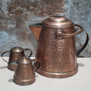 May include: A set of three copper teapots with hammered surfaces. The largest teapot has a lid and a handle. The two smaller teapots have lids and handles.