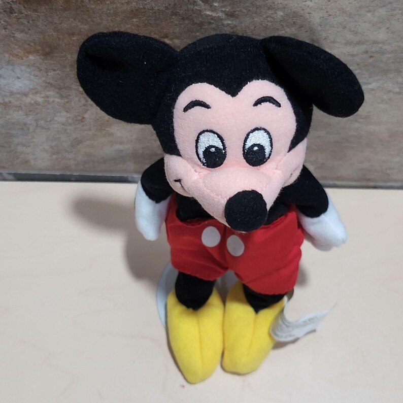 May include: A plush Mickey Mouse toy. The toy features black ears, a black head, a pink face, and a black nose. It wears red shorts with white buttons, yellow shoes, and white gloves. The toy is a classic Disney character.