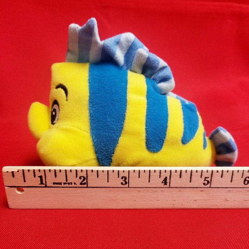 K&ouml;nnte beinhalten: Ein gelbes und blaues Pl&uuml;schtier in Form eines Fisches mit einer blau-wei&szlig; gestreiften Flosse. Das Spielzeug ist etwa 13 cm lang, gemessen mit einem Holzlineal. Der Fisch hat ein l&auml;chelndes Gesicht und ist vor rotem Hintergrund platziert.