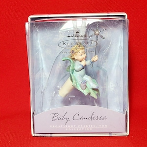 May include: A Hallmark Keepsake Ornament featuring a baby fairy with a blue dress and green wings, holding a silver wand with a star on top. The ornament is in a clear plastic box with a white background and the text "Baby Candessa Frostlight Fairies Too Collection" on the bottom.