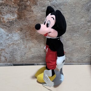 May include: A Mickey Mouse plush toy, featuring a black head and ears, a pink muzzle, and a black nose. The toy wears a red pair of shorts, white gloves, and yellow shoes. The plush is mounted on a silver stand.