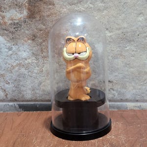 May include: A small orange Garfield figurine with a grumpy expression and crossed arms, standing on a black circular pedestal base. The character is enclosed within a clear, dome-shaped glass display cover that sits securely on the black base.