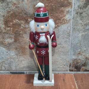 May include: A decorative wooden nutcracker in a skiing theme. The nutcracker is painted burgundy with white snowflake accents and a white beard. It wears a red hat with green trim and white pom-pom. It holds skis and a ski pole.