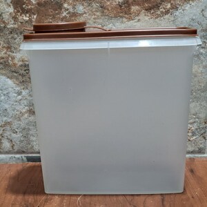 Puede incluir: Un recipiente de plástico transparente con una tapa marrón. El recipiente es alto y rectangular.