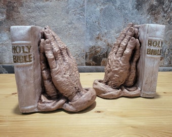 May include: A pair of bookends shaped like praying hands, each holding a book with the words "Holy Bible" printed on the cover. The bookends are made of a light brown material and have a rustic look.