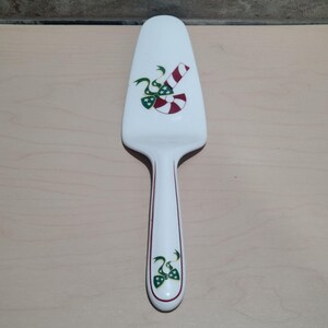 May include: White ceramic pie server with a candy cane and bow design. The server has a long handle with a red stripe down the centre. The candy cane is red and white, and the bows are green and red.