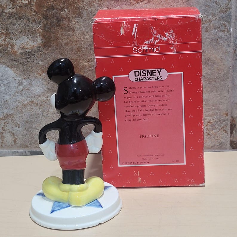 Puede incluir: Una figurita de cer&aacute;mica de Mickey Mouse, de pie con las manos en las caderas. La figurita est&aacute; pintada en negro, rojo y amarillo, y est&aacute; colocada sobre una base blanca con un dise&ntilde;o de estrella azul. La figurita est&aacute; empaquetada en una caja roja con las palabras "Schmid Disney Characters Figurine" impresas en ella.