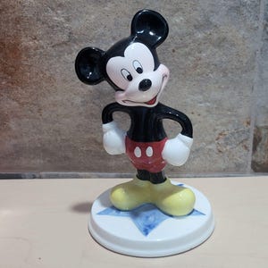 Puede incluir: Una figurita de cer&aacute;mica de Mickey Mouse de pie sobre una base azul en forma de estrella. Mickey lleva sus pantalones cortos rojos caracter&iacute;sticos con botones negros y zapatos amarillos.