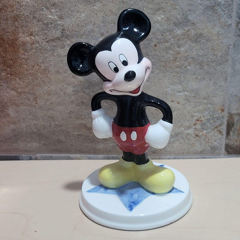 Puede incluir: Una figurita de cer&aacute;mica de Mickey Mouse de pie sobre una base azul en forma de estrella. Mickey lleva sus pantalones cortos rojos caracter&iacute;sticos con botones negros y zapatos amarillos.
