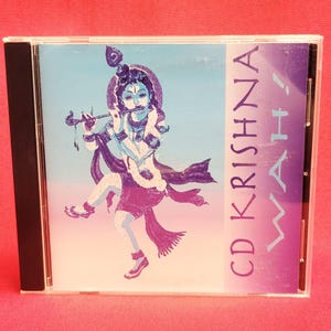 May include: A CD case featuring a vibrant illustration of a deity playing a flute, with the text "CD KRISHNA WAH!". The artwork is in shades of purple, blue, and white against a gradient background. The CD case is in a clear plastic case.
