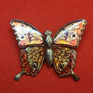 Puede incluir: Un broche de mariposa de tono dorado con alas de purpurina naranja y morada. La mariposa tiene un cuerpo y antenas detalladas.