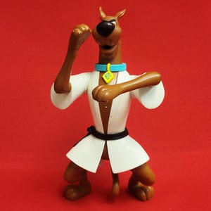 Puede incluir: Figura de Scooby-Doo marrón con uniforme de karate, con un gi blanco, cinturón negro y collar azul con colgante amarillo. La figura posa en posición de karate sobre un fondo rojo.