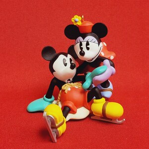 Peut inclure: Une figurine d'ornement de Mickey Mouse et Minnie Mouse de style vintage. Mickey porte un pantalon rouge et des patins à glace bleus. Minnie porte une robe à pois rouges et blancs et des patins à glace bleus. Ils sourient tous les deux et se tiennent la main.
