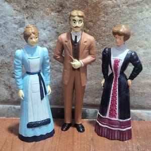 May include: Three vintage figurines of people in period clothing. A man in a brown suit and two women in dresses. One woman wears a light blue dress with a white apron, and the other wears a black dress with red accents.