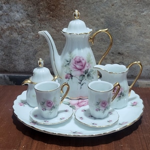 Puede incluir: Un juego de té de porcelana blanca que incluye una tetera, un azucarero con tapa, una lechera y dos tazas con sus platos a juego, dispuestos sobre una bandeja redonda. Cada pieza está decorada con motivos de rosas rosas y bordes dorados.