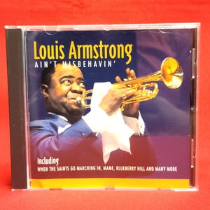 Puede incluir: Caja de CD con Louis Armstrong tocando la trompeta. El título del álbum es "Ain't Misbehavin'" en texto amarillo. La portada incluye los nombres de canciones como "When The Saints Go Marching In" y "Blueberry Hill". El fondo es azul oscuro.