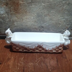 May include: White ceramic rectangular dish with a basket weave design and two white cat figurines on the sides. The dish has a white interior and a decorative fabric with cherries.