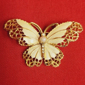 May include: A gold-tone butterfly brooch with white enamel wings and a pearl-like center stone. The wings are detailed with a delicate filigree design.
