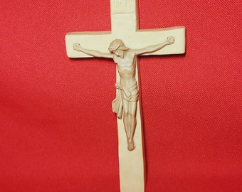 May include: A cream-colored crucifix with a detailed depiction of Jesus Christ. The cross is rectangular with a slightly curved design. The background is a solid red color.