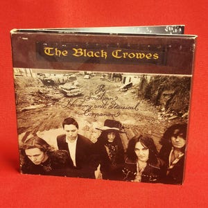 May include: A CD case with the band name "The Black Crowes" in gold lettering. The album cover features a sepia-toned image of a group of people and vintage cars. The album title is "The Southern Harmony and Musical Companion."