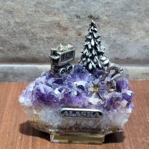 Peut inclure: Un objet décoratif composé d'un amas de cristaux d'améthyste violette surmonté d'une figurine en métal argenté représentant un mineur, un sapin et un wagonnet rempli de minerai. Une plaque argentée portant le mot ALASKA est fixée à l'avant.
