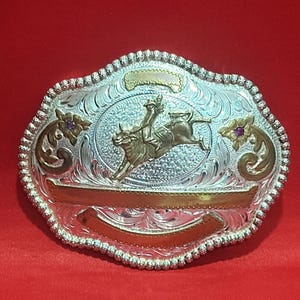 May include: A silver-toned belt buckle featuring an oval centre with a gold-toned bull rider figure. The buckle has ornate scrollwork engravings, gold-toned accents, and two small purple stones set within floral motifs on the left and right sides.