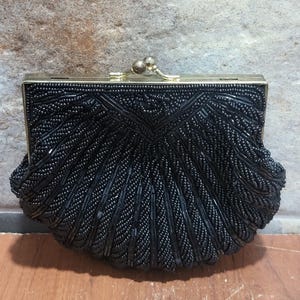 May include: Black beaded clutch with a gold-toned clasp. The clutch features a fan-like design with intricate beadwork. The rectangular frame contrasts with the rounded bottom, creating a vintage aesthetic. A stylish accessory for special occasions.