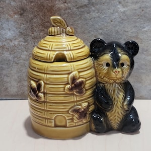 May include: A ceramic honey pot shaped like a beehive with a bear figurine. The beehive is yellow with brown bees and a lid. The bear is black and tan, sitting upright next to the beehive.