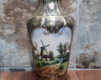 May include: A brown and gold ceramic vase with a painted scene of a windmill and a village. The vase has a decorative gold trim and a white background.
