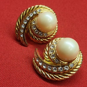 May include: A pair of gold tone earrings with a faux pearl and clear rhinestones. The earrings are in the shape of a swirl.