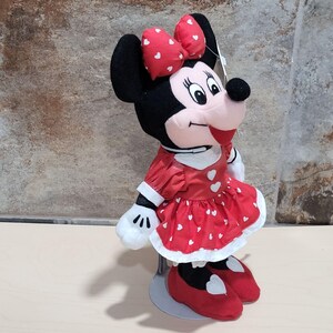 K&ouml;nnte beinhalten: Minnie Mouse Pl&uuml;schpuppe, mit einem roten Kleid mit wei&szlig;en Herzakzenten, einer passenden Schleife und roten Schuhen. Die Puppe hat schwarze Ohren und ein rosa Gesicht. Die Puppe steht auf einem silbernen St&auml;nder.