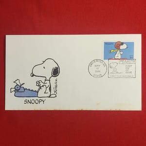 以下が含まれることがあります： スヌーピーのイラスト、タイプライター、そして「SNOOPY」の文字が描かれた白い封筒。飛行士の格好をしたスヌーピーが描かれた切手。封筒には、2001年5月17日、カリフォルニア州サンタローザの消印が押されています。