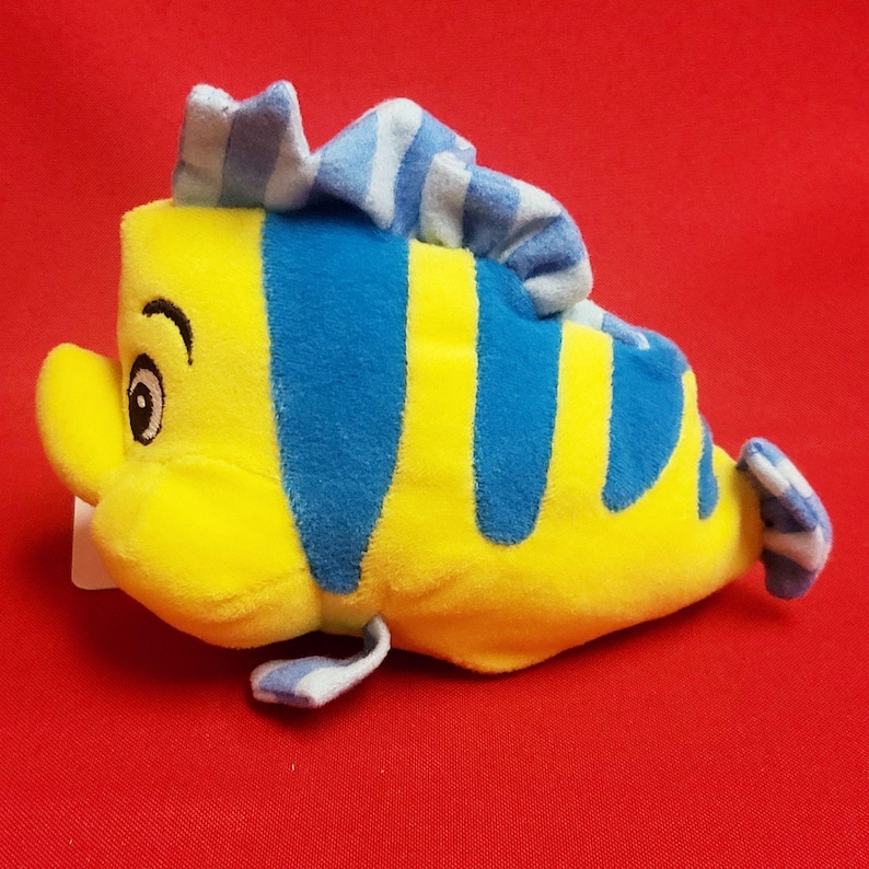 K&ouml;nnte beinhalten: Ein gelb-blaues Pl&uuml;schtier in Form eines Fisches mit einem Cartoon-Design. Der Fisch hat blaue Streifen, eine blau-wei&szlig; gestreifte Flosse und ein blau-wei&szlig; gestreiftes Kopfst&uuml;ck. Er hat ein l&auml;chelndes Gesicht mit einem Auge. Der Hintergrund ist rot.