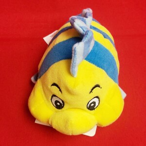 K&ouml;nnte beinhalten: Ein gelbes und blaues Pl&uuml;schfischspielzeug mit einem l&auml;chelnden Gesicht und gro&szlig;en Augen. Das Spielzeug hat blaue Streifen und eine blaue Flosse. Der Hintergrund ist einfarbig rot.