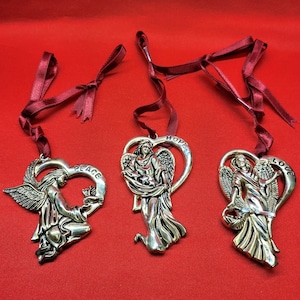 May include: Three silver angel ornaments with red ribbon hangers. Each angel is holding a heart-shaped frame with the words "Peace", "Hope", and "Love" inscribed on them.