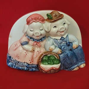 Könnte beinhalten: Ein Keramik-Serviettenhalter mit zwei Schweinefiguren. Das weibliche Schwein trägt ein rosa Kleid mit blauen Verzierungen und einer roten Schleife. Das männliche Schwein trägt eine blaue Latzhose und einen braunen Hut. Sie halten einen Korb mit grünen Äpfeln.