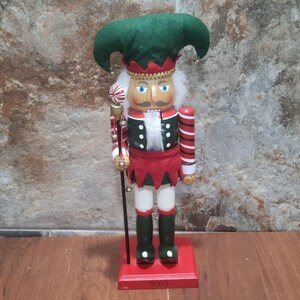 May include: A wooden nutcracker with a green and red jester costume, holding a candy cane staff. The nutcracker has a white beard and blue eyes. The base of the nutcracker is red and has the year 2005 printed on it.