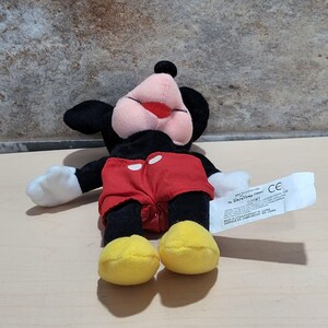 May include: A plush Mickey Mouse toy. The toy has a black body, ears, and head, with a pink face and red shorts. The hands are white, and the shoes are yellow. A white tag is attached to the toy.