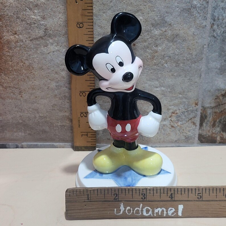 Puede incluir: Una figurita de cer&aacute;mica de Mickey Mouse de pie sobre una base azul en forma de estrella. Mickey lleva sus pantalones cortos rojos caracter&iacute;sticos con dos botones negros y zapatos amarillos. Est&aacute; en posici&oacute;n con las manos en las caderas y tiene una sonrisa traviesa en la cara.