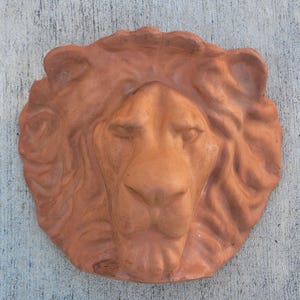 Puede incluir: Escultura de cabeza de león de terracota con rasgos faciales detallados y una melena texturizada. La escultura es de un color marrón anaranjado cálido y terroso, probablemente para decoración exterior o de jardín.