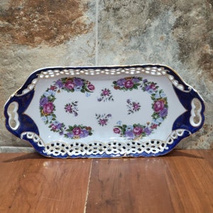 Puede incluir: Una bandeja de servir de porcelana blanca con ribetes azules y dorados. La bandeja tiene un diseño floral y un borde de encaje. Tiene dos asas a los lados.