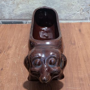 Puede incluir: Una maceta de cerámica marrón con forma de perro salchicha. La maceta tiene forma de perro con una gran abertura para plantas. El perro tiene un acabado brillante y está sobre una superficie de madera. Es un artículo decorativo único.