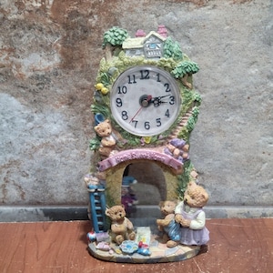 May include: A decorative clock with a whimsical design. The clock face is white with black numbers and hands. The clock is surrounded by a scene with teddy bears, a house, and the words "Home Sweet Home" on a banner. The clock is approximately 20 cm tall.