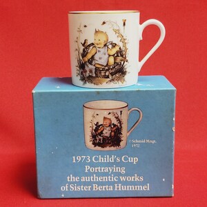 Può includere: Una tazza in ceramica bianca con un bordo dorato e un'immagine di un bambino seduto in un cesto con uccelli e fiori. La tazza è su una scatola blu con il testo "1973 Child's Cup Portraying the authentic works of Sister Berta Hummel" e "Schmid Mngt. 1972".