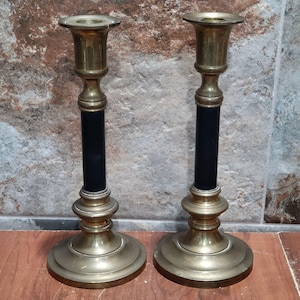 May include: Two antique brass candlesticks with black detailing. The candlesticks feature a traditional design with a broad base, a central black section, and a flared top for candles. The brass has a warm, aged patina.