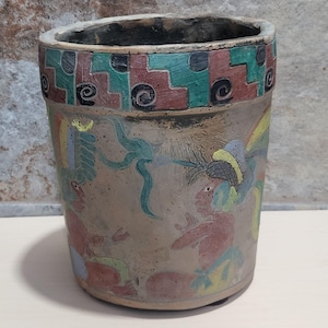 May include: A cylindrical, hand-painted ceramic pot with a beige base and a colorful geometric border. The pot features detailed illustrations of figures in red, yellow, blue, and green. The design suggests an ancient or tribal theme.