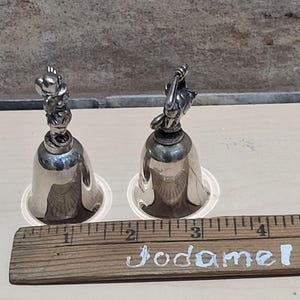 Pu&ograve; includere: Due campanelli color argento con figurine di animali sulla parte superiore. I campanelli sono appoggiati su basi di plastica trasparente. Un righello di legno con la scritta "Jodamel" &egrave; in primo piano. I campanelli sono alti circa 7.6 cm.